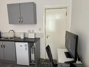 Una cocina compacta con refrigerador, lavamanos y microondas. Se ve una puerta con manija y un televisor sobre un mueble.