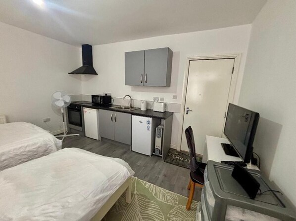 Una habitación de hotel compacta con dos camas individuales, kitchenette, televisor y un escritorio pequeño.