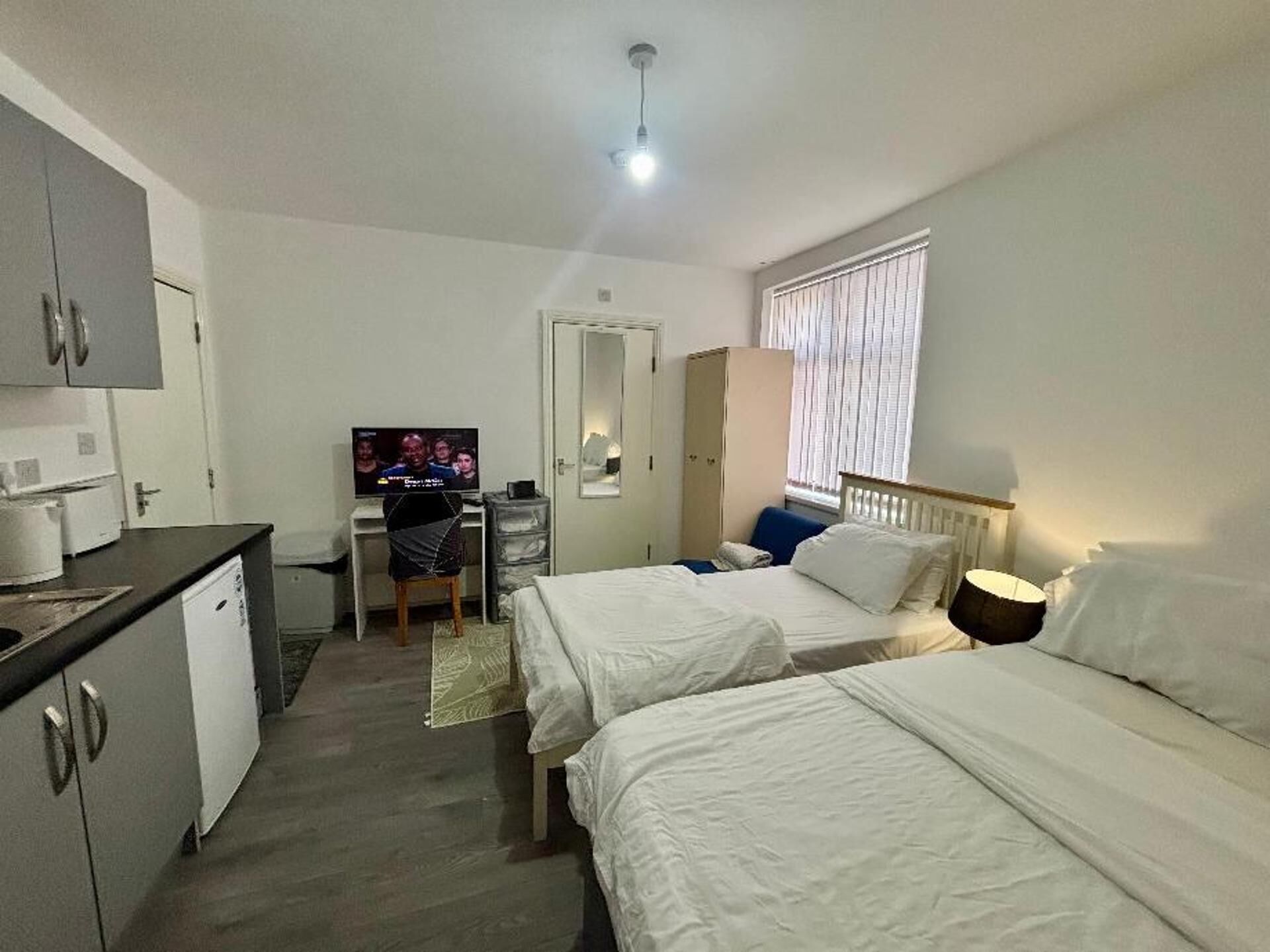 Quarto de hotel com duas camas, uma escrivaninha com televisão, um guarda-roupa e uma janela com persianas.