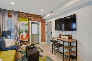 A modern living room with a brick accent wall, a flat-screen TV mounted on the wall, a yellow sofa, a coffee table, and a dining area with a wooden table and chairs. - Comfy 1 Bedroom Apartment! Salt Lake City. Close to UofU (Salt Lake City)