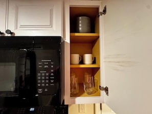 A kitchen with a microwave and a cabinet containing mugs and glasses.