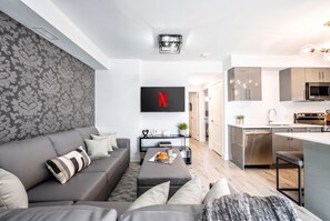 A modern living room with a grey sofa, a flat-screen TV mounted on the wall, and a kitchen area with stainless steel appliances. - Lux 2BR Retreat With Parking, 5 Mins to Falls and Sleeps 6 Guests! (Niagara Falls)