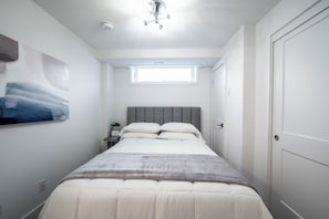 Quarto com cama, mesa de cabeceira, relógio na parede, janela e porta.
