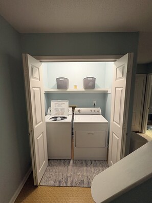 A laundry room with a washing machine and dryer, a shelf with baskets, and a patterned rug.