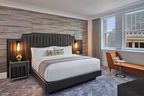 Une chambre d’hôtel moderne avec un grand lit, une table de chevet avec une lampe, un coin salon avec un fauteuil et une vue sur la ville par la fenêtre.