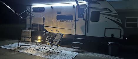 A well-lit RV with the brand 'Passport' on the side, a small table with a lamp, two chairs, and a concrete floor.