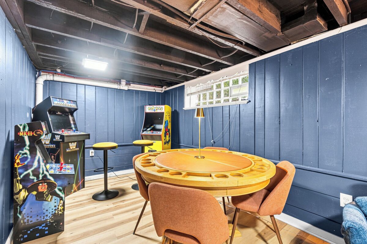 A room with a round wooden table and orange chairs, arcade machines, and a window with blinds.