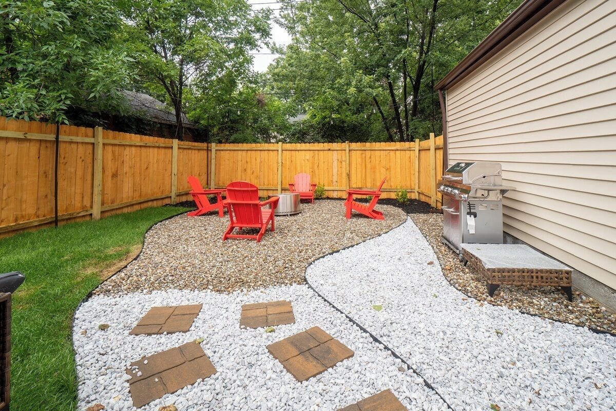 A backyard with a wooden fence, red chairs, a grill, and a paved area with stepping stones.
