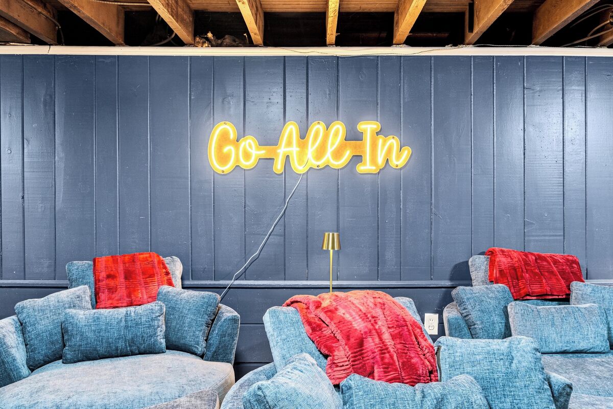 A room with a neon sign that reads 'Go All In' on the wall, a sofa with a red blanket, and a lamp.