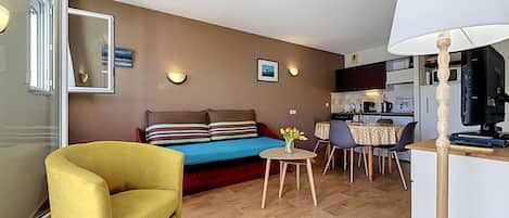 Un espacio habitable compacto con sofá, un sillón amarillo, una mesa de comedor y una kitchenette.