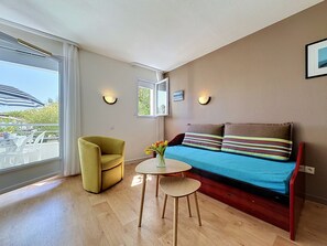 Ein Zimmer mit einer Couch, einem gelben Sessel, einem kleinen runden Tisch mit Blumenvase und einem Fenster mit Vorhängen.