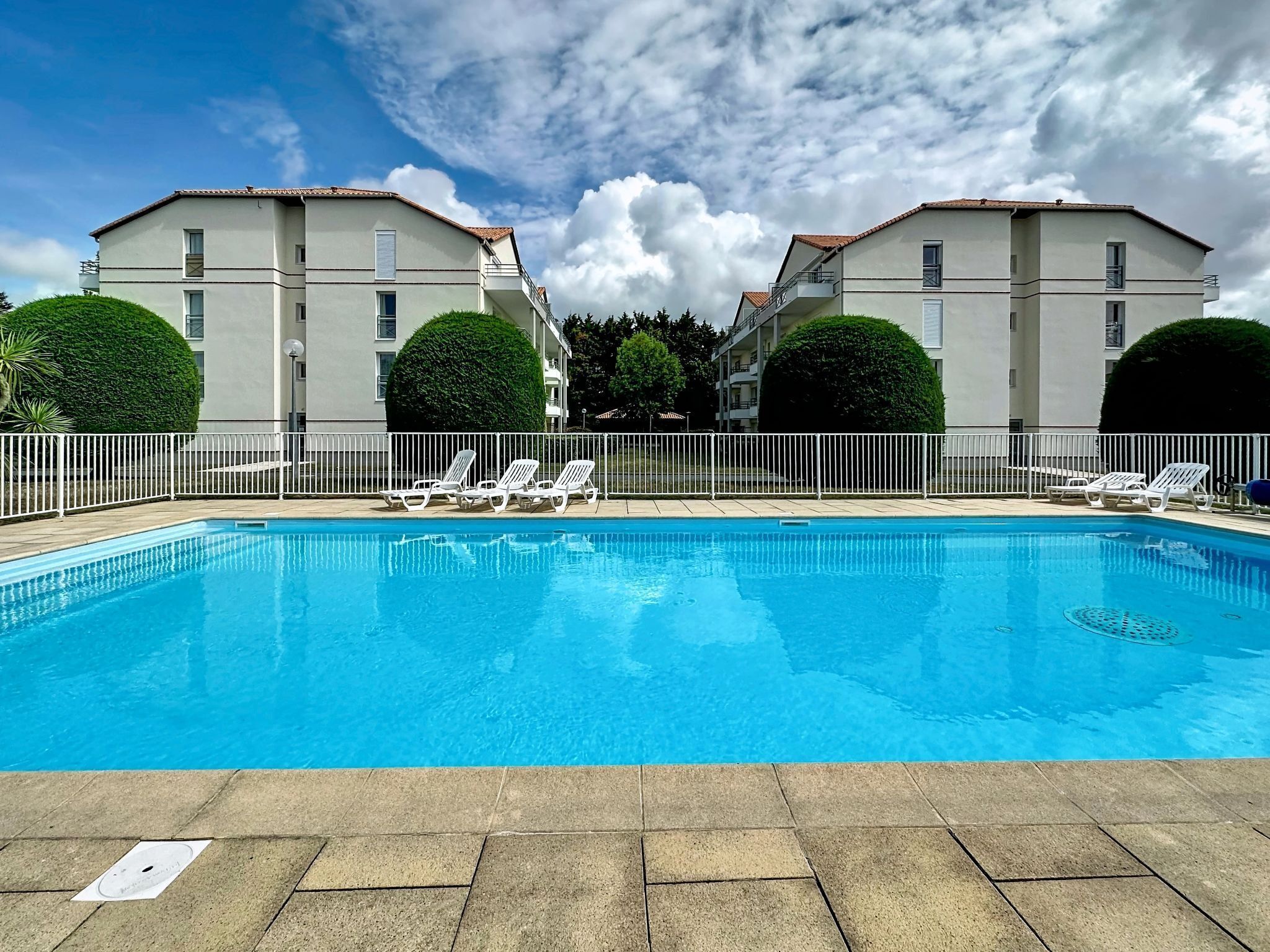 A swimming pool with lounge chairs, surrounded by a white fence, and adjacent to a building with balconies.