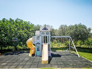 Un parc infantil amb tobogan i cadira enfilada, envoltat d'arbres.