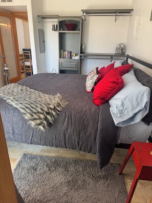 Ein Schlafzimmer mit einem Bett, einem roten und weißen Kopfkissen, einer grauen Decke und einem kleinen roten Tisch.