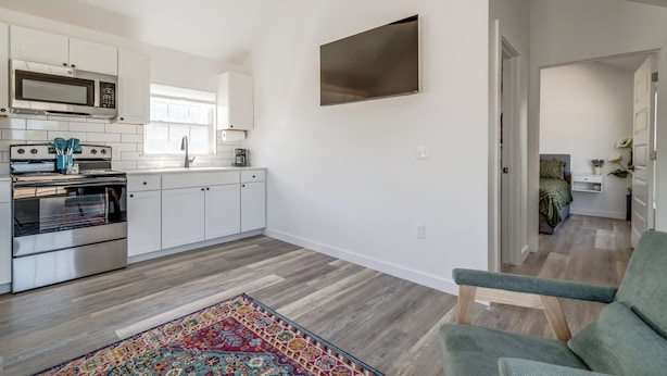 A modern kitchen with white cabinets, a microwave, and a large window. There is a colorful rug on the floor and a green chair in the living area. - 66 Cottage on Historic Rt 66 -Starlit Stone (El Reno)