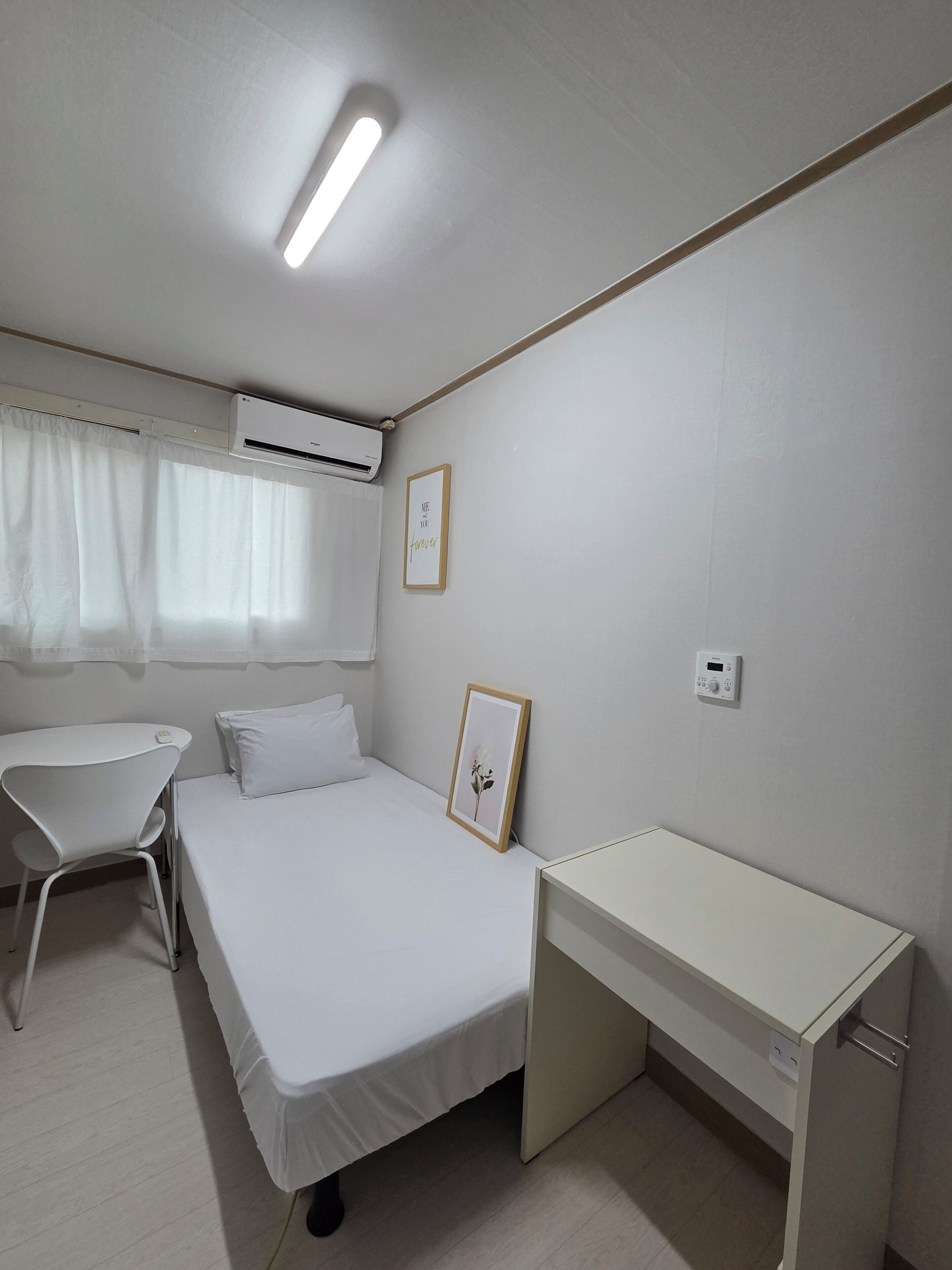 Una stanza piccola e pulita con un letto, una scrivania, una sedia, un condizionatore d'aria e una finestra con tende.