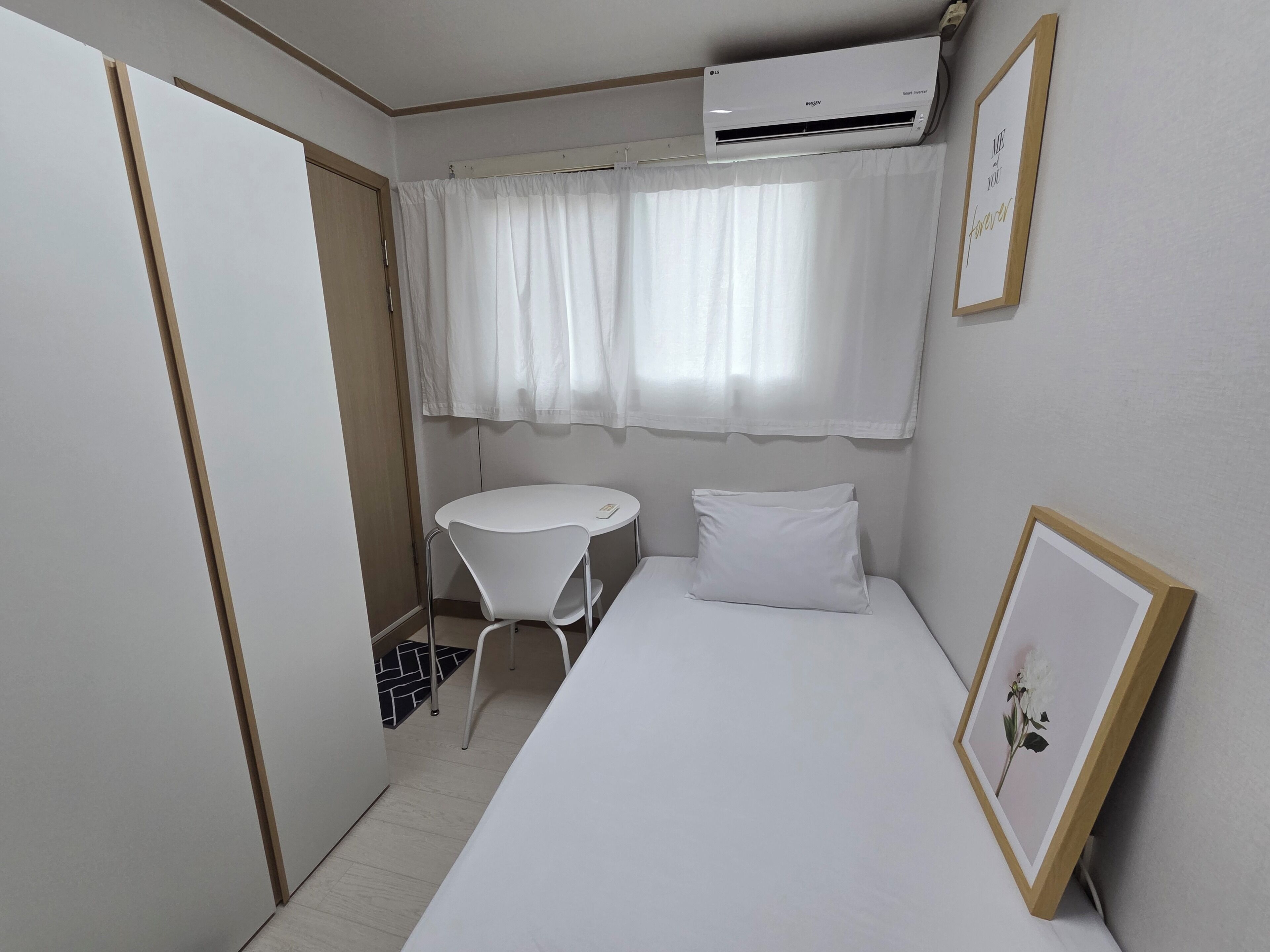 Una stanza piccola e ordinata con un letto, un tavolo e una sedia. Un condizionatore d'aria è montato sulla parete sopra il letto.