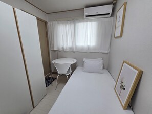 Ein kleiner, ordentlicher Raum mit einem Bett, einem Tisch und einem Stuhl. Eine Klimaanlage ist an der Wand über dem Bett angebracht.