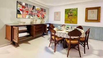 A dining area with a table set for four, surrounded by chairs. There is a wooden cabinet with drawers and a shelf, and a large abstract painting on the wall.