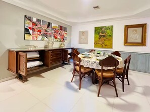 A dining area with a table set for four, surrounded by chairs. There is a wooden cabinet with drawers and a shelf, and a large abstract painting on the wall. - Dúplex Premium - Piscinas, Jacuzzi & Sauna (Santa Cruz de la Sierra)