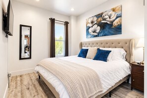 A bedroom with a large bed, a flat-screen TV mounted on the wall, a window with curtains, and a painting above the bed. - Salt Lake City Comfort! 1 Bed 1 Bath (Salt Lake City)