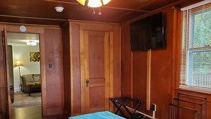 A room with a bed, a television mounted on the wall, a window with blinds, and a door leading to another room. - Goodyear Lake Boathouse - charming historic cottage overlooking the water (Oneonta)