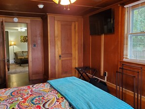 A room with a bed, a television mounted on the wall, a window with blinds, and a door leading to another room. - Goodyear Lake Boathouse - charming historic cottage overlooking the water (Oneonta)