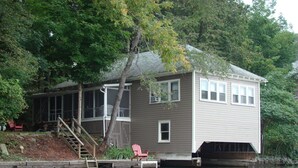 A house with a dock extending into a calm body of water, surrounded by trees. - Goodyear Lake Boathouse - charming historic cottage overlooking the water (Oneonta)