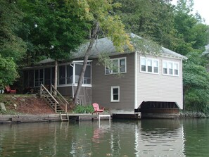 A house with a dock extending into a calm body of water, surrounded by trees. - Goodyear Lake Boathouse - charming historic cottage overlooking the water (Oneonta)