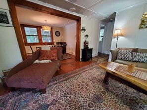 A living room with a sofa, a coffee table, a patterned rug, a fireplace, and a dining area with a table and chairs. - Goodyear Lake Boathouse - charming historic cottage overlooking the water (Oneonta)