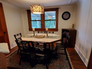 A dining area with a table, chairs, a patterned tablecloth, a candle holder, a window with curtains, a clock on the wall, and a bookshelf. - Goodyear Lake Boathouse - charming historic cottage overlooking the water (Oneonta)