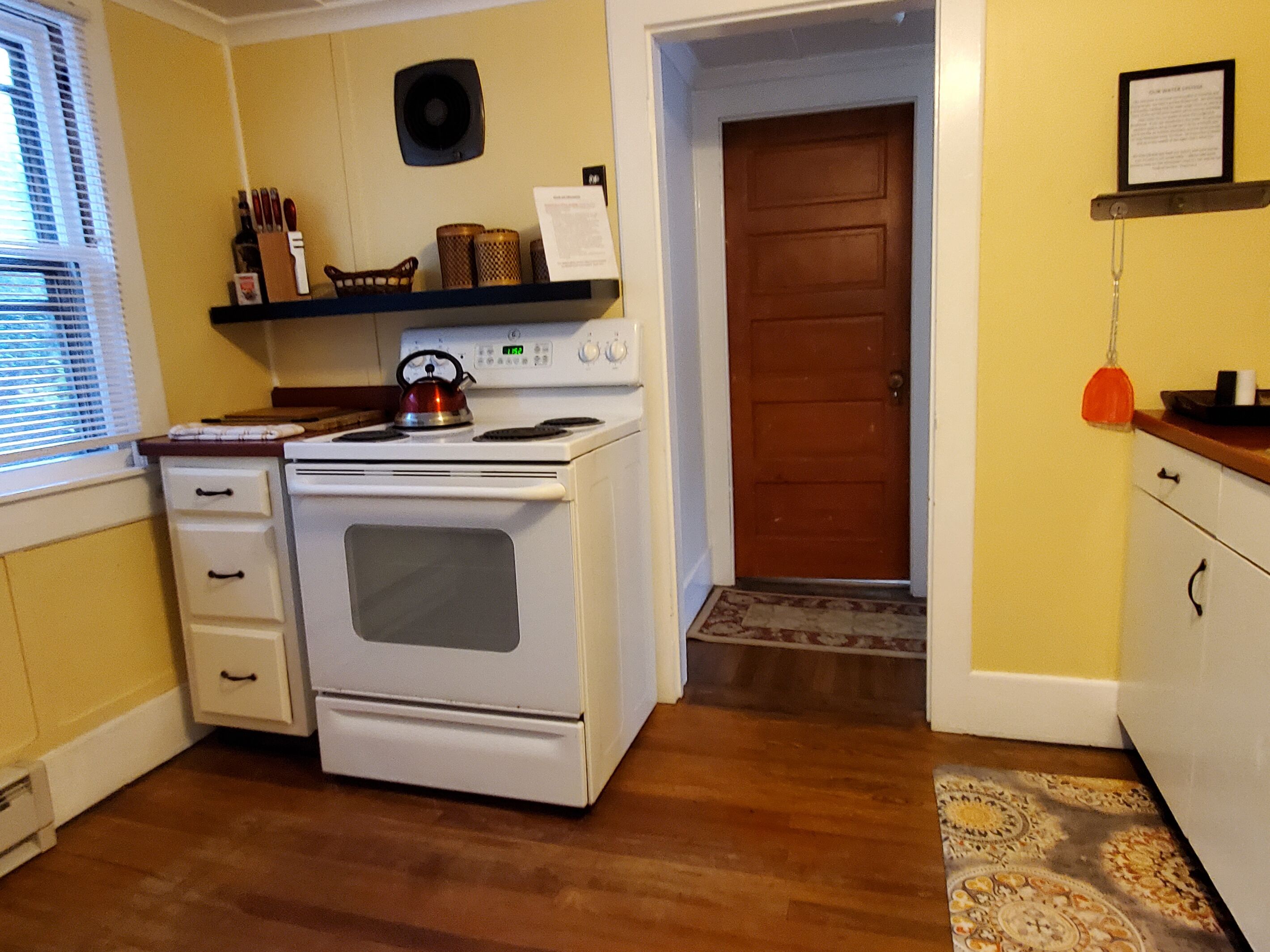 A kitchen with a stove, oven, and a kettle. There is a wooden floor, a window with blinds, and a door leading to another room.