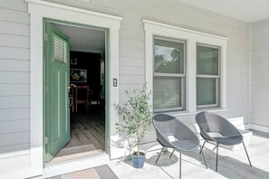 Una entrada de casa con puerta verde, dos ventanas, una planta en maceta y dos sillas afuera.