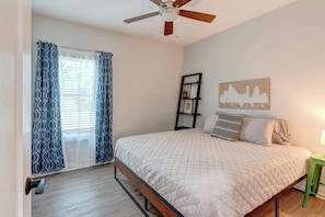 Quarto com cama, ventoinha no teto, janela com cortinas, quadro na parede e mesa de cabeceira.