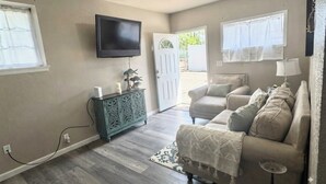 Una sala de estar con un sofá color beige, un televisor montado en la pared y una puerta que da al exterior.