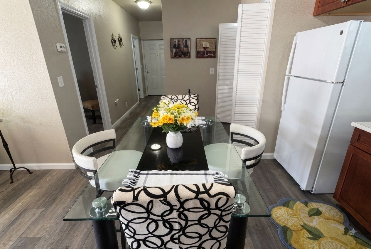 Un coin repas avec une table en verre, des chaises, un vase de fleurs et un réfrigérateur.