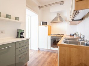 Une cuisine compacte équipée d’un four encastré, d’un four à micro-ondes et d’une cuisinière à gaz. Elle comprend des armoires en bois et un évier en acier inoxydable.
