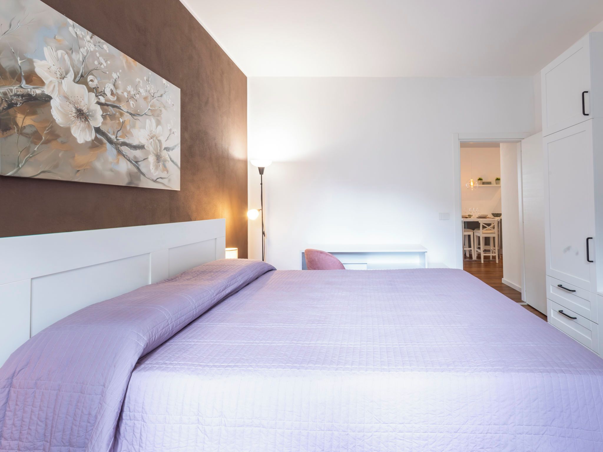 A bedroom with a large bed, a painting on the wall, and a view into the kitchen.