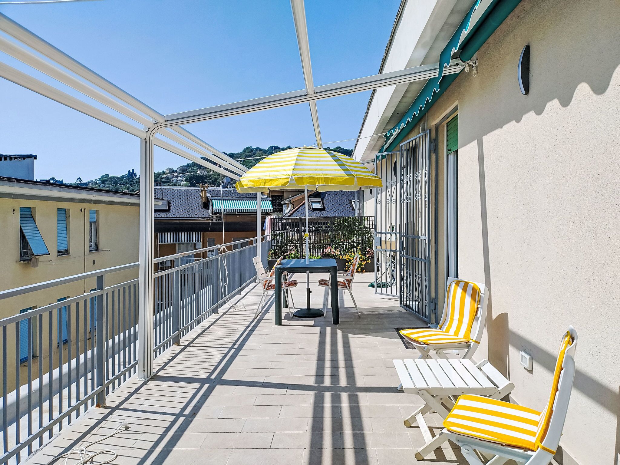 A balcony with a table and chairs, a yellow striped umbrella, and a view of buildings in the background.