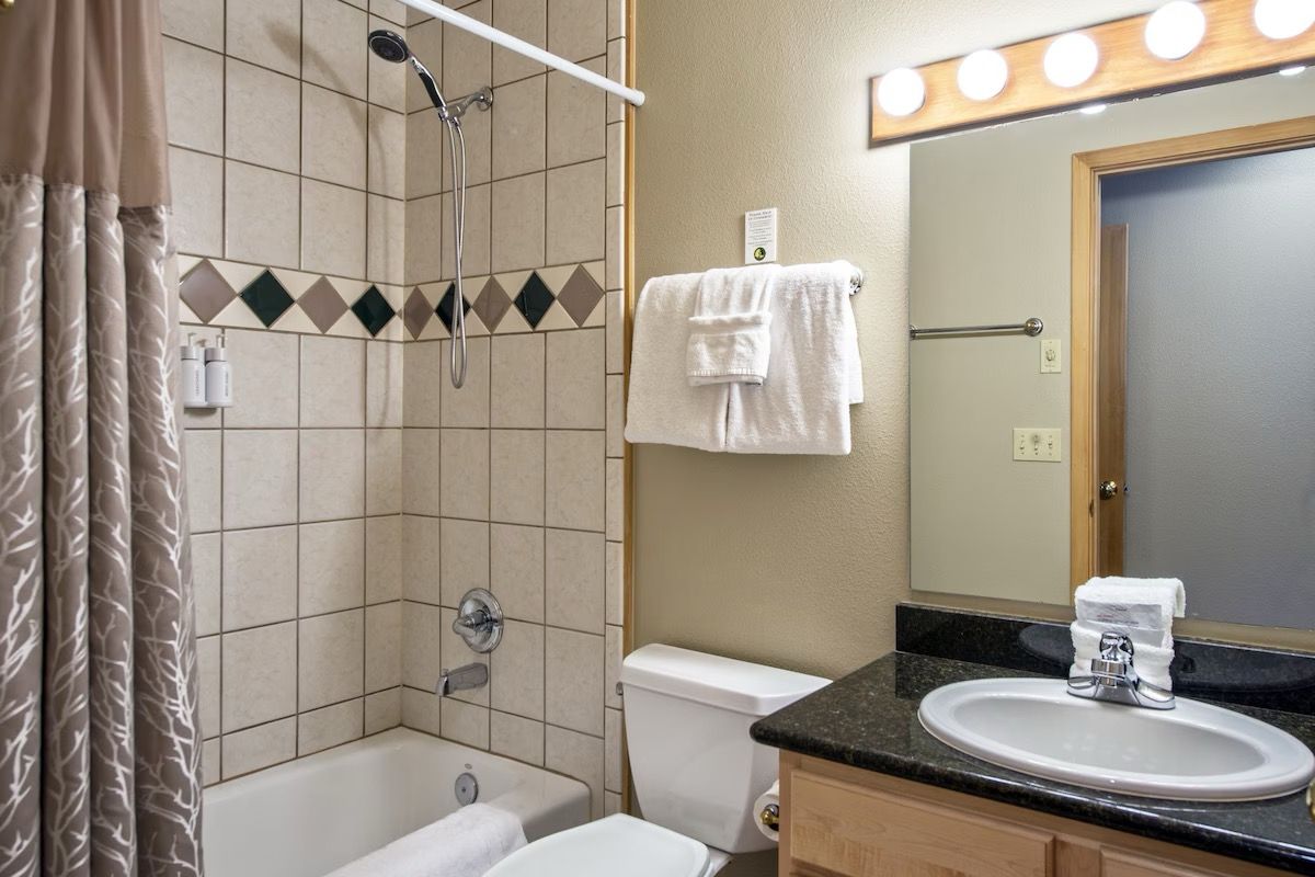 A bathroom with a bathtub, toilet, and sink. There is a showerhead, a towel rack, and a mirror with lights above it.