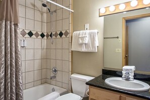 Un baño con bañera, inodoro y lavabo. Hay un cabezal de ducha, un perchero y un espejo con luces arriba.