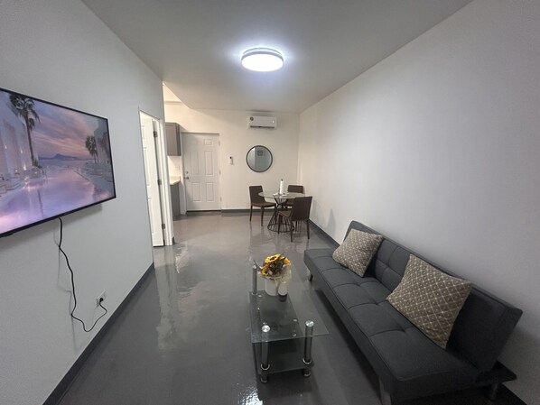 Una sala de estar moderna con un televisor de pantalla plana, una mesa de centro de vidrio con flores, un sofá con cojines estampados y un comedor con sillas y una mesa.