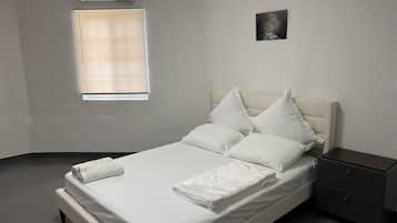 Un lit bien fait, avec du linge de lit blanc et des oreillers, une table de chevet avec un tiroir, une petite lampe de chevet et une fenêtre avec des stores.