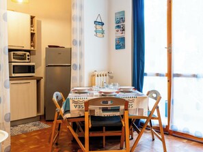 A compact kitchen with a refrigerator, microwave, and toaster, a dining table set with plates and glasses, and a view through a window with a blue curtain. - Margherita by Interhome (Santo Stefano al Mare)