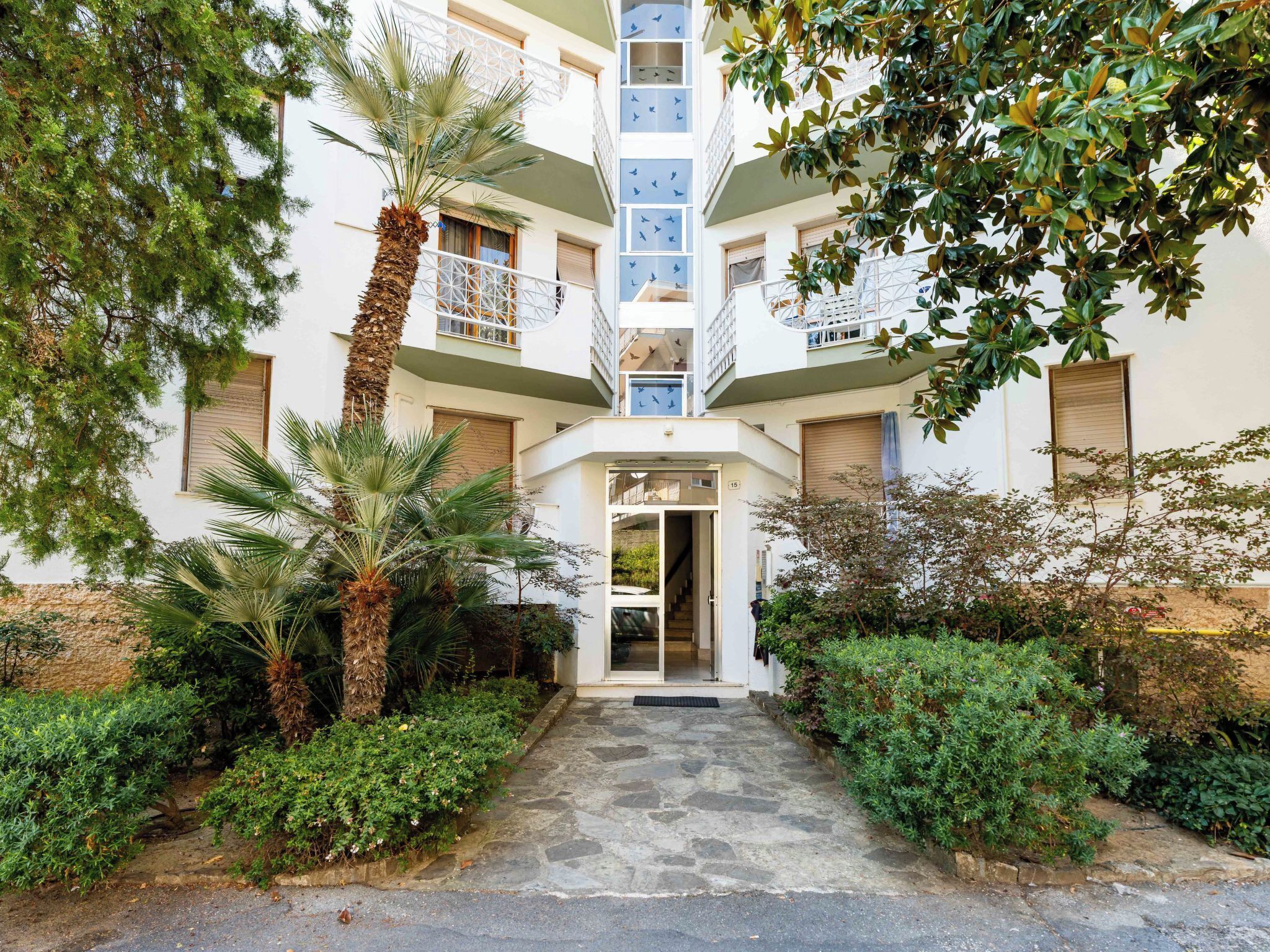 A multi-story building with a central entrance, surrounded by greenery and palm trees.