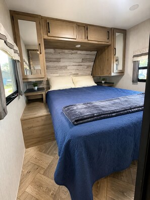 A bedroom with a bed, wooden cabinets, a small table, and a window. - 2-bedroom recreational vehicle in beautiful Shingleton with AC (Shingleton)
