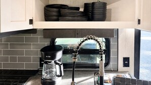 A compact kitchen with a coffee maker, a window, and a shelf with stacked plates. - 2-bedroom recreational vehicle in beautiful Shingleton with AC (Shingleton)