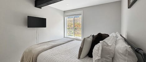 Quarto com cama, televisão de tela plana na parede e janela com vista para árvores.