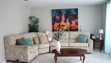 A living room with a beige sectional sofa, a round wooden coffee table, a large painting of palm trees, and a potted plant.