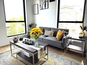 Una sala de estar moderna con un sofá gris, una mesa de centro con un jarrón de girasoles y vistas a edificios a través de grandes ventanales.